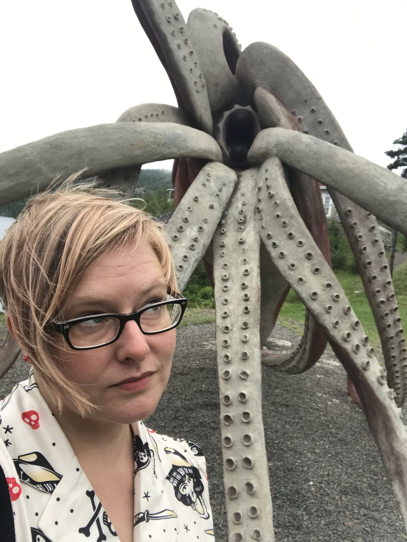 Picture of me with short blonde hair and black glasses, looking sideways at the maw of a statue of the Giant Squid in Glover's Harbour, Newfoundland.